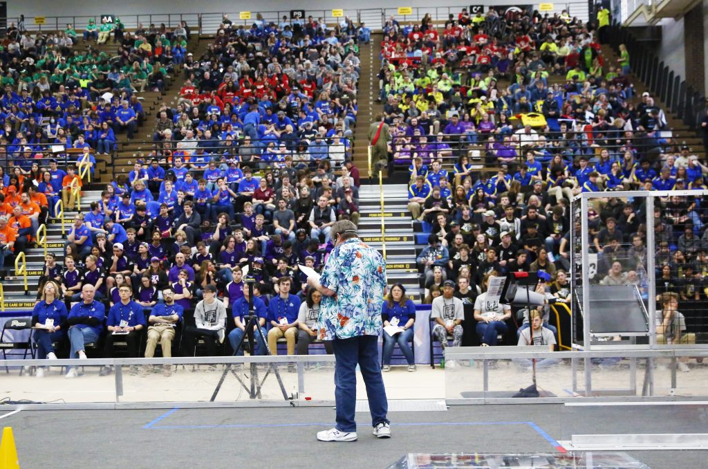 Man in blue shirt standing at podium in front of crowd of people.