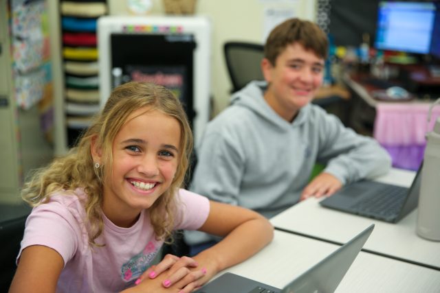 girl in pink sitting in classroom smiling with boy in grey sweatshirt sitting behind her also smiling