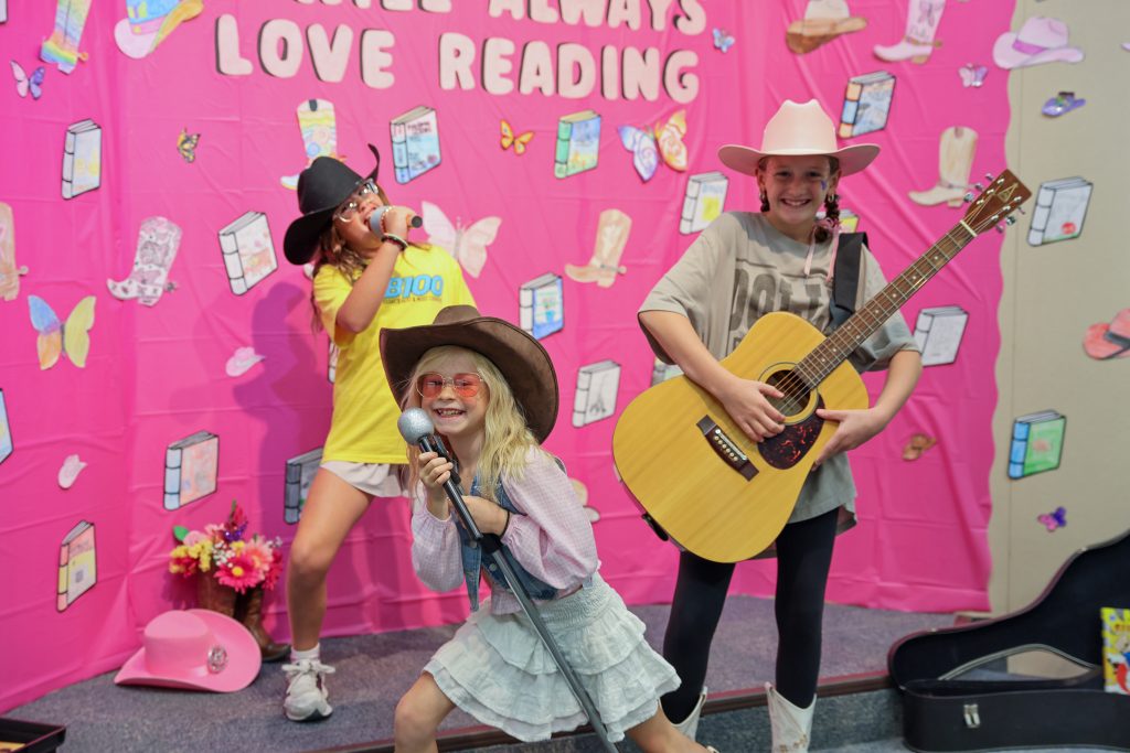Three children dressed in cowboy outfits, holding guitars and singing on a stage.