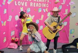 Three children dressed in cowboy outfits, holding guitars and singing on a stage.